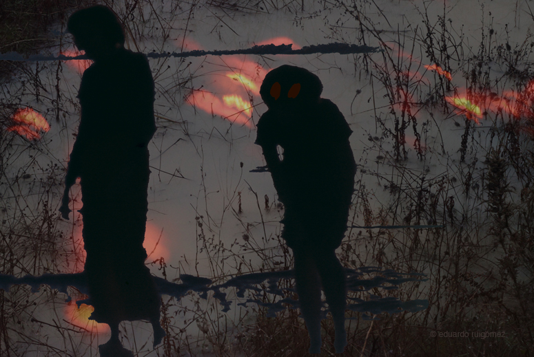 Dos mujeres en sombra en una ciénaga. Al fondo hay unas luces rojas desperdigadas. En la silueta de la derecha se pueden ver dos manchas rojo oscuro a la altura de los ojos.